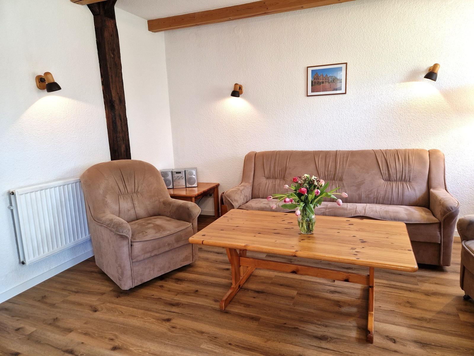 Wohnzimmer mit Sofa, Sessel, Holztisch und Blumen. Holzdielenboden und Wandlampen.
