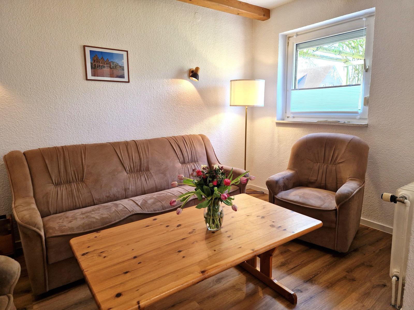 Wohnzimmer mit Sofa, Sessel und Holztisch. Fenster und Wandlampen sichtbar.