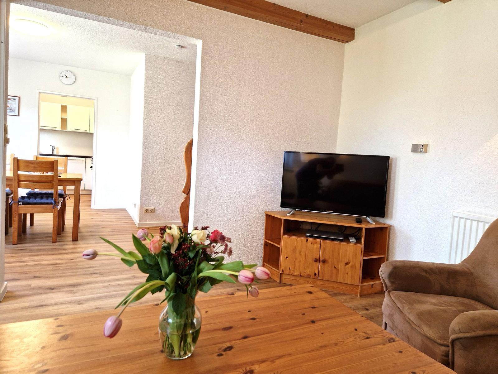 Wohnzimmer mit Fernseher, Holztisch und Blumen. Küche und Essbereich im Hintergrund.