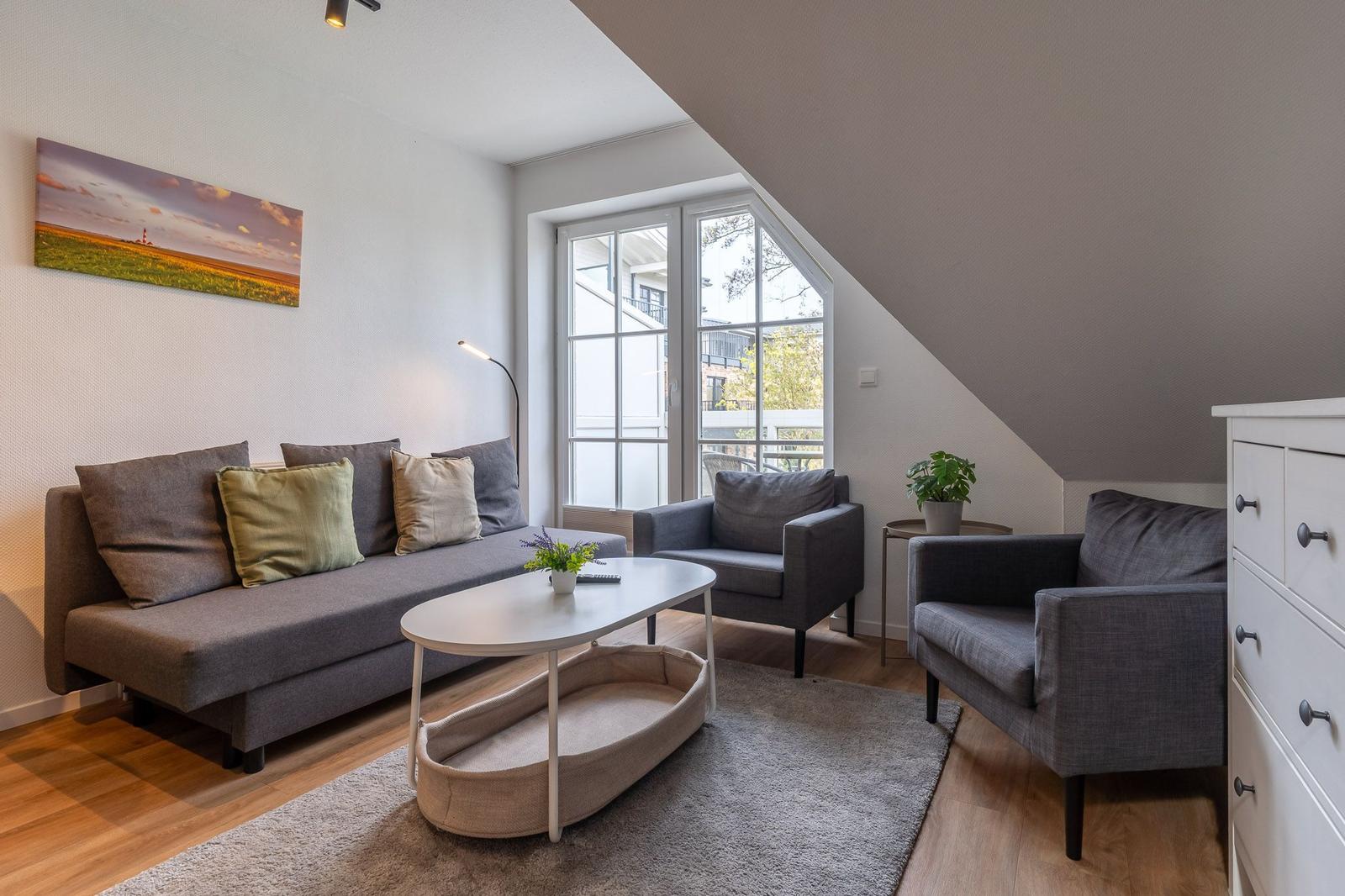 Wohnzimmer mit Sofa, zwei Sesseln, Tisch und Fenster. Wandbild und Pflanzen sind sichtbar.