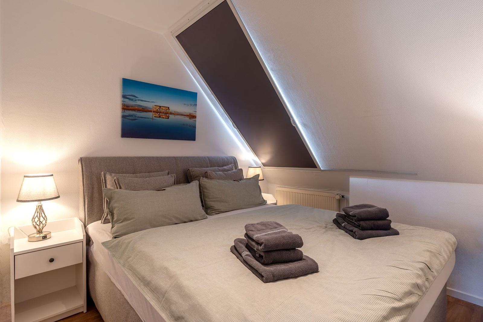 Schlafzimmer mit Dachschrägen, Bett mit Kissen und Handtüchern, Nachttisch mit Lampe und Wandbild.