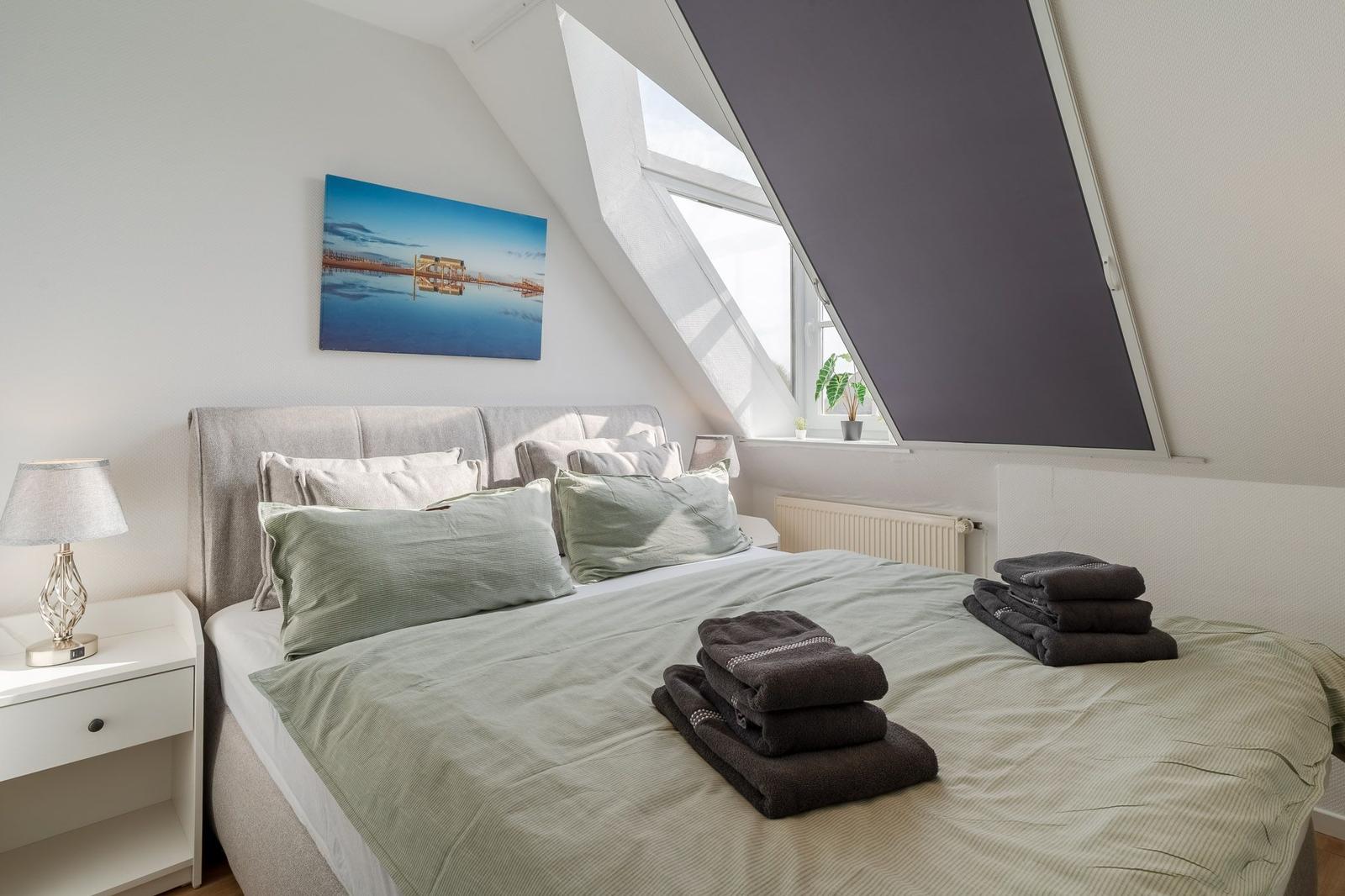 Schlafzimmer mit Dachschrägen, Bett mit Kissen und Handtüchern, Fenster und Wandbild.