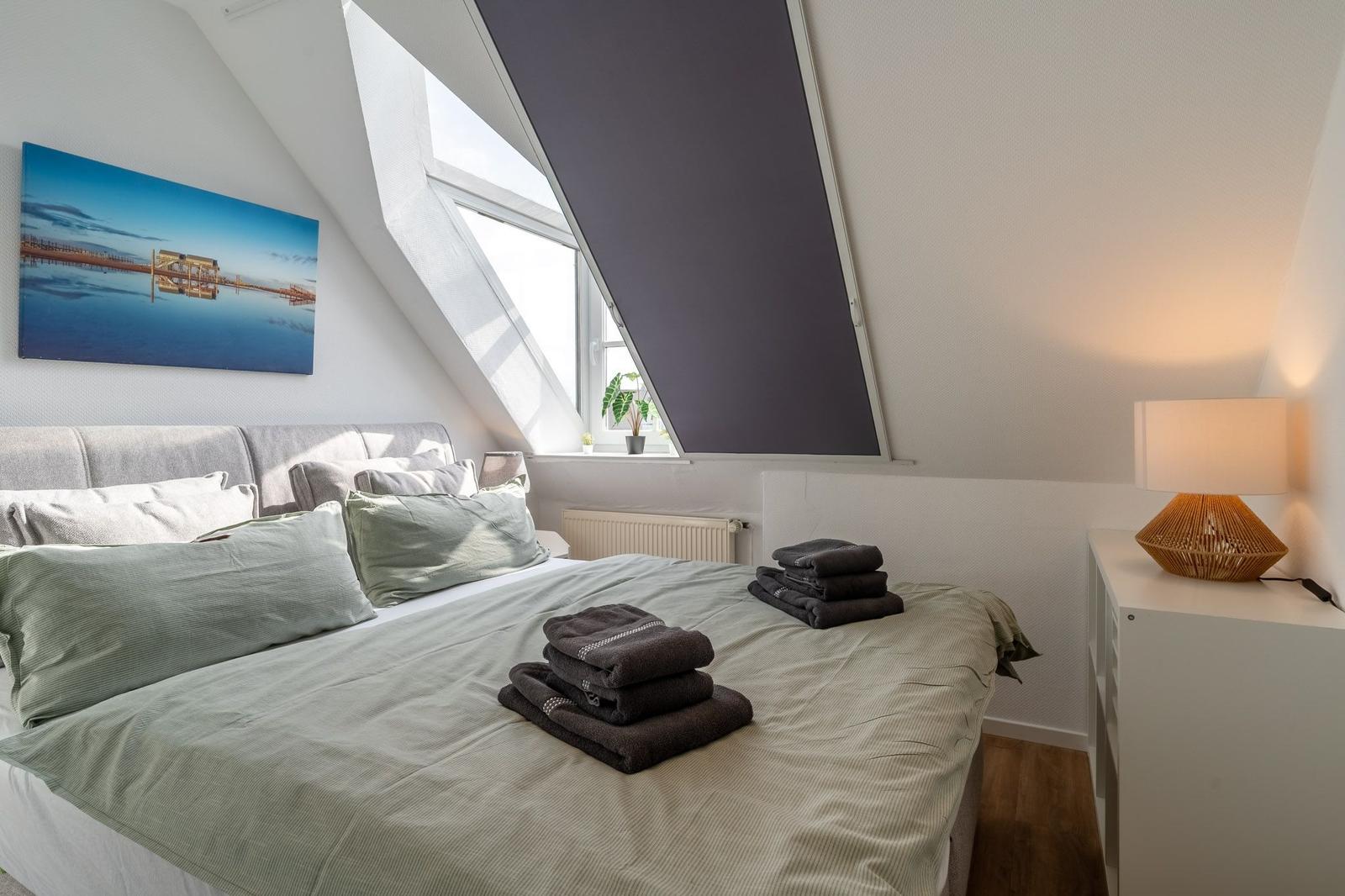 Schlafzimmer mit Dachschrägen, Bett mit Handtüchern, Fenster und Wandbild.