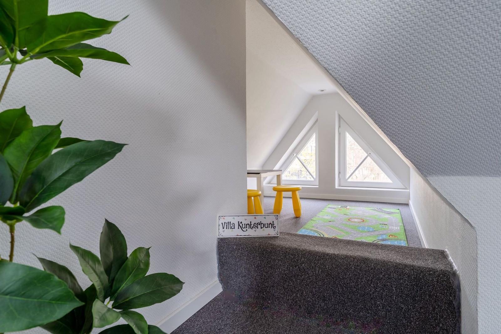 Kinderzimmer mit Spielteppich, gelben Hockern und Dreiecksfenstern unter Dachschrägen.