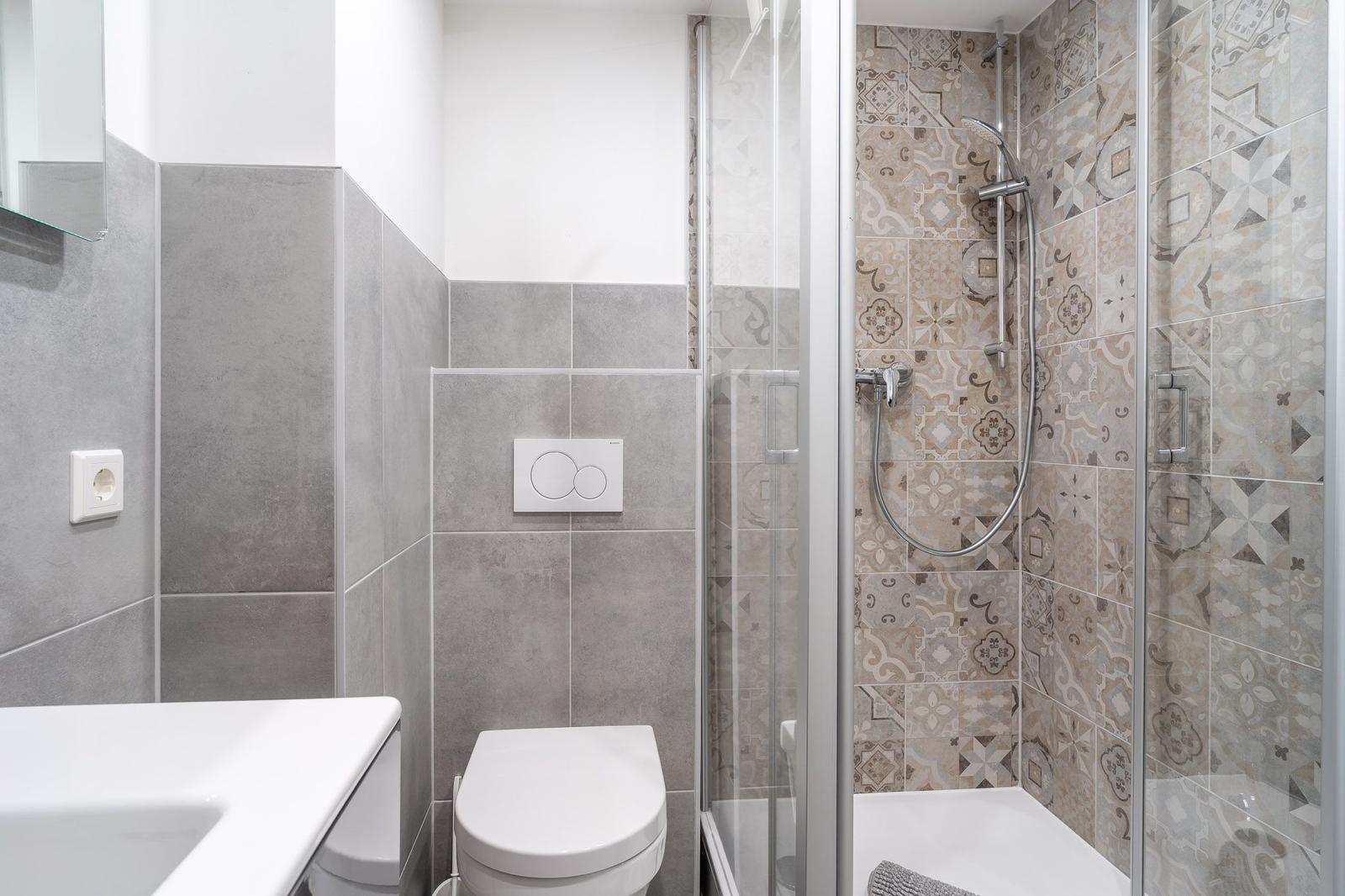 Badezimmer mit Dusche, Toilette und Waschbecken. Wandfliesen in Grau und Muster.
