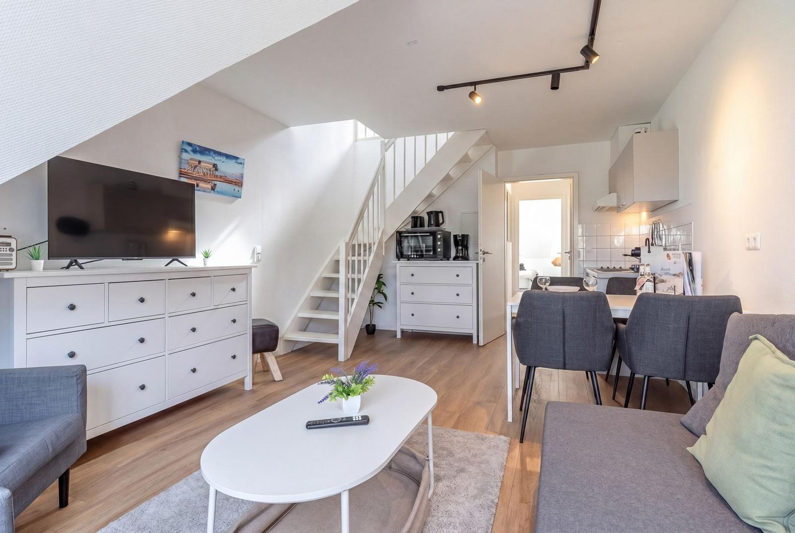 Wohnbereich mit Sofa, Couchtisch und TV. Treppe führt zur Küche. Holzboden und weiße Wände.
