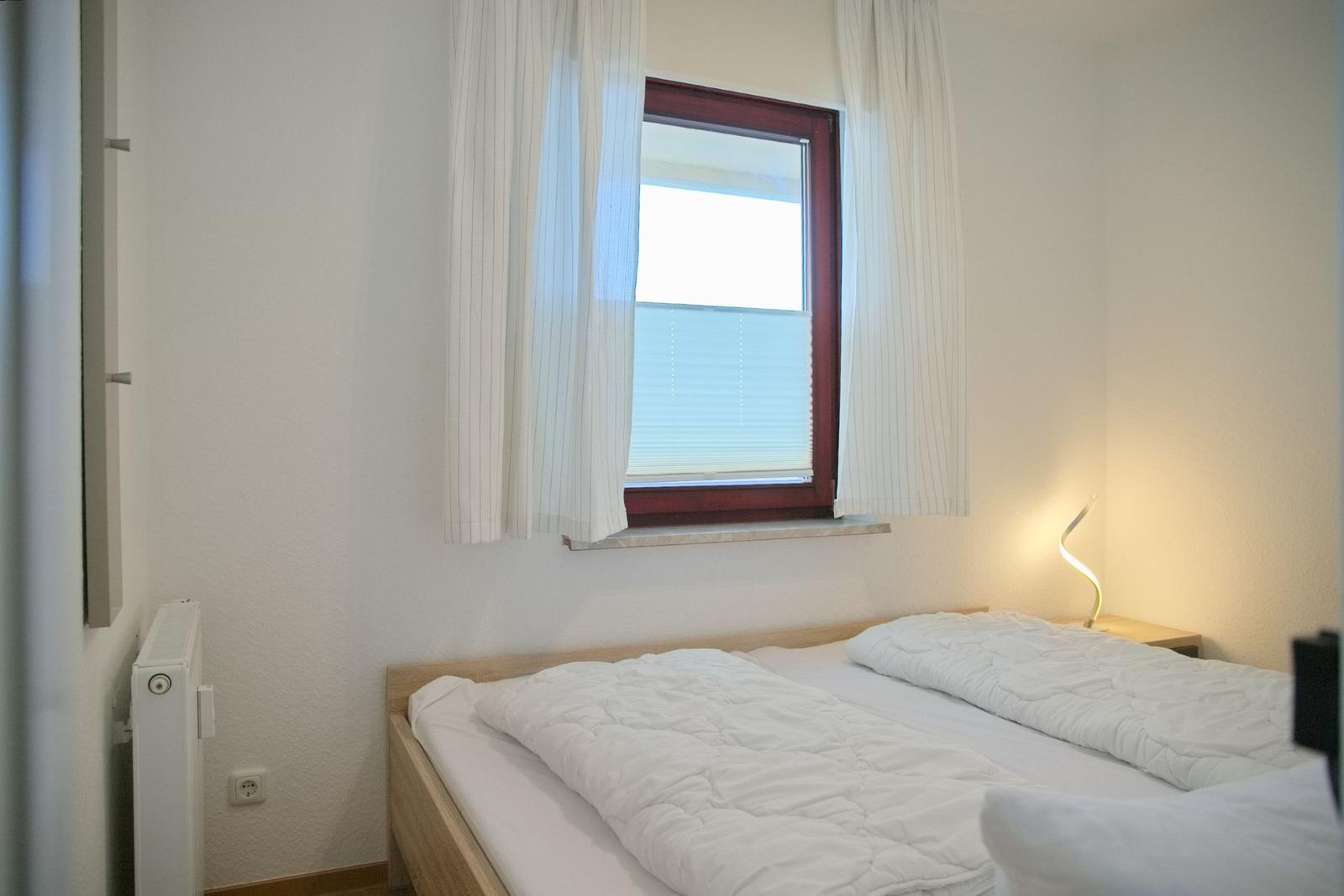 Ein Schlafzimmer mit einem Bett, einem Fenster mit weißen Vorhängen und einer Stehlampe.