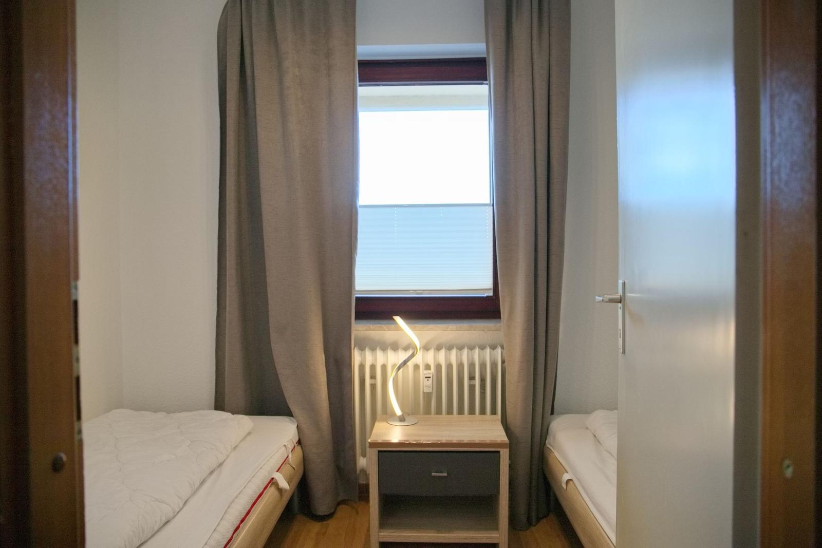 Zwei Einzelbetten mit Nachttisch und Lampe unter Fenster mit Jalousie und Vorhängen.