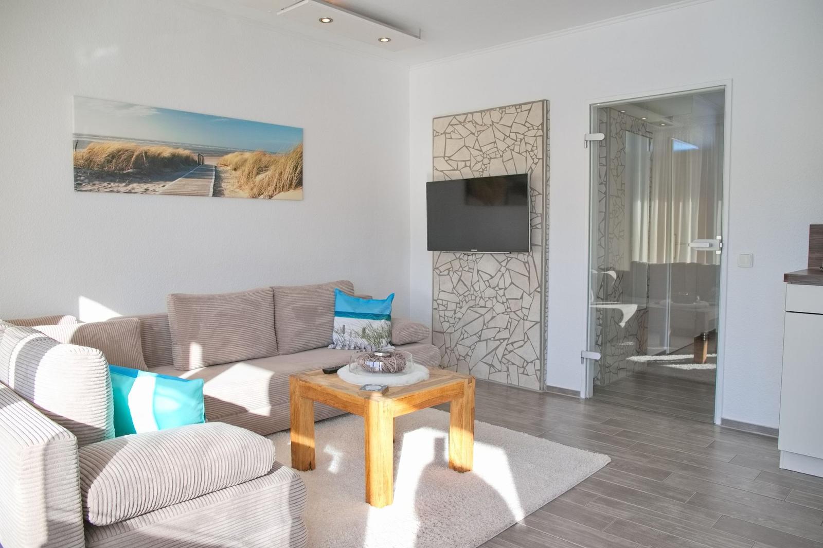 Wohnzimmer mit Sofa, Couchtisch und Fernseher an Wand. Durchgang zur Badewanne.