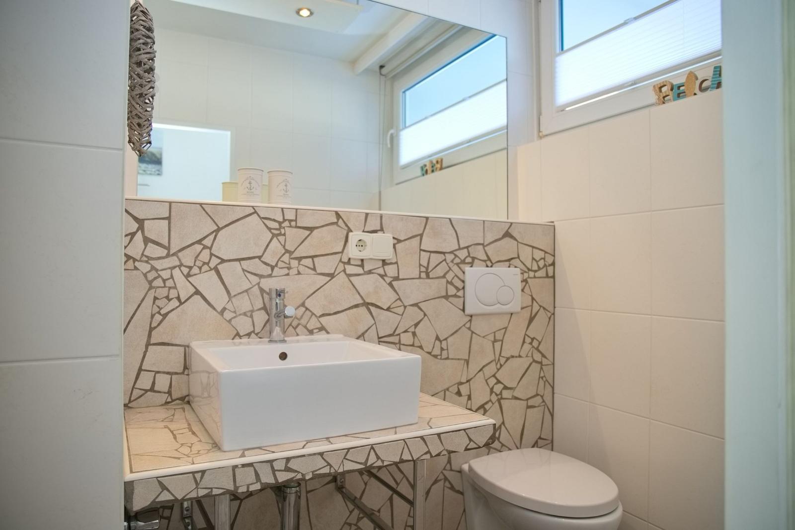 Badezimmer mit Waschbecken, Spiegel und Toilette. Wandverkleidung aus Mosaikfliesen.
