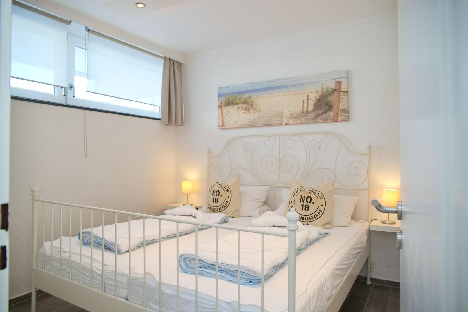 Schlafzimmer mit weißem Metallbett, Fenster und Strandbild an der Wand.