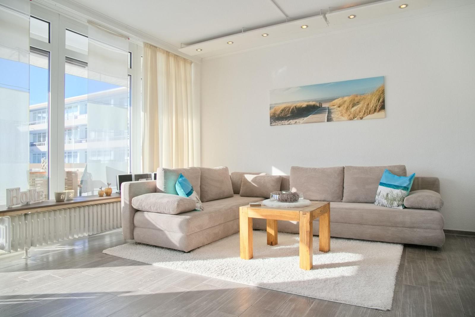 Wohnzimmer mit Ecksofa, Holztisch und Wandbild. Große Fenster mit Blick nach draußen.