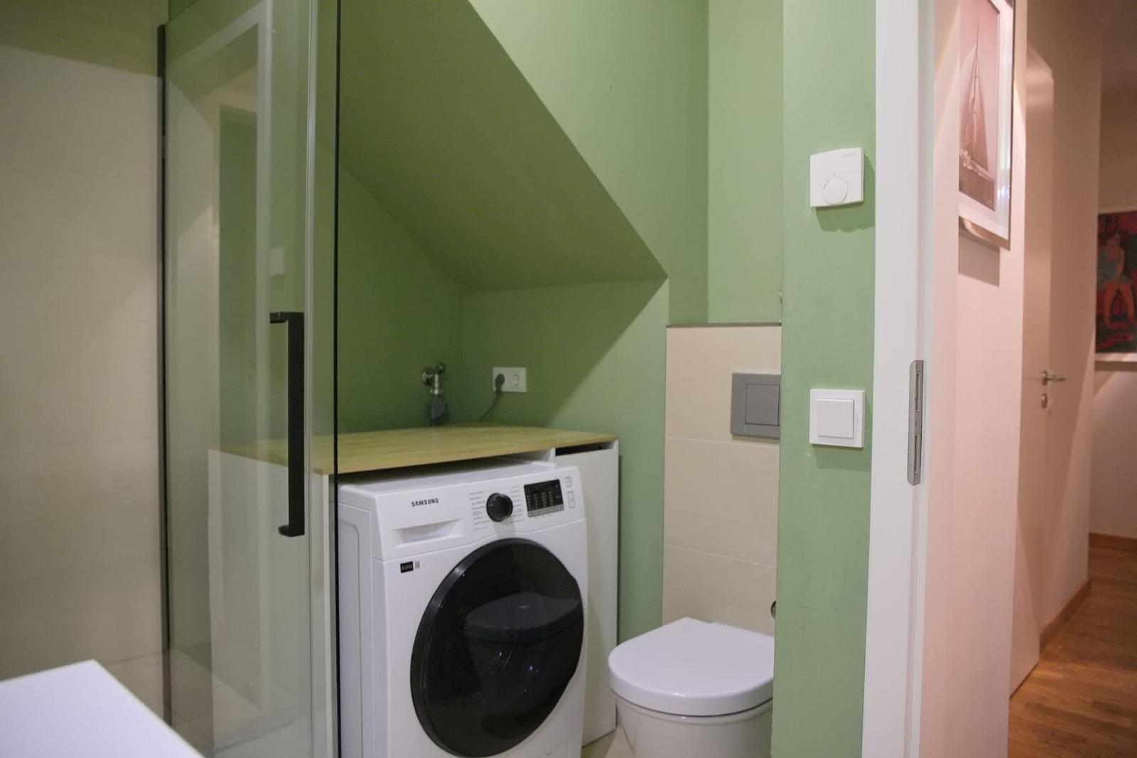 Badezimmer mit Waschmaschine, Toilette und Dusche mit Glaswand.