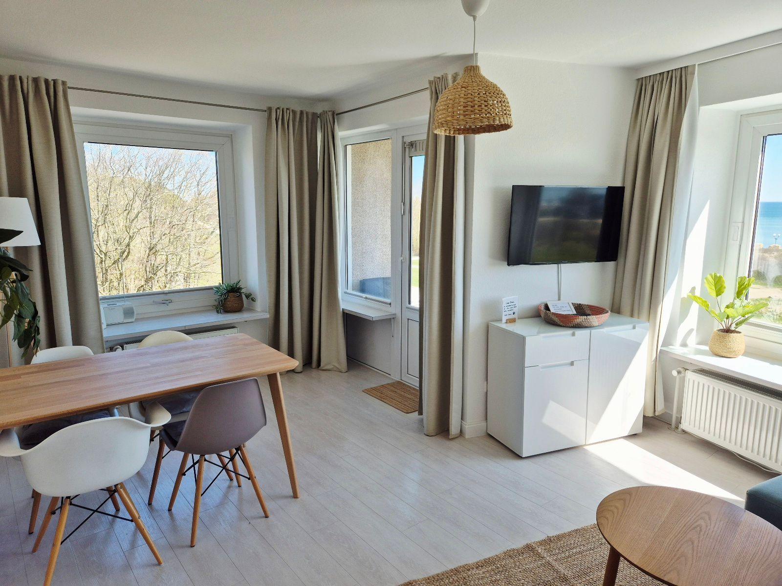 Wohn- und Essbereich mit Blick auf Meer und Wald. Holztisch, TV und Pflanzen.
