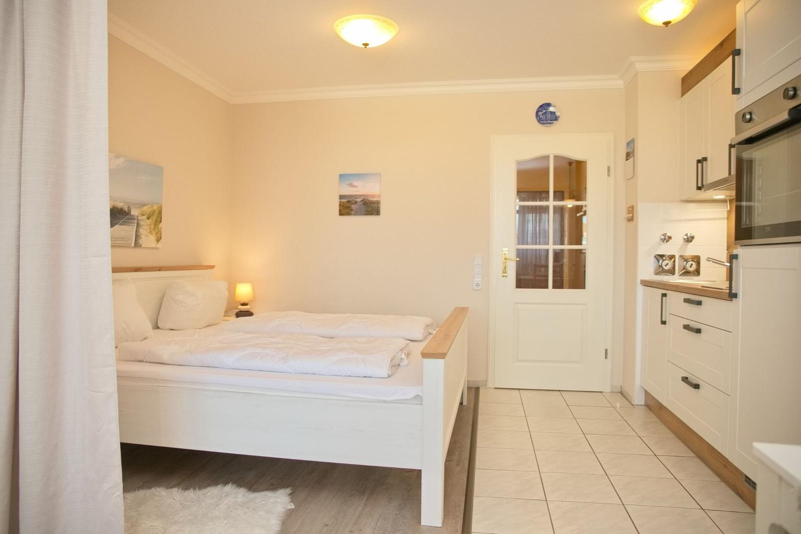 Schlafzimmer mit Doppelbett, Tür und Küchenbereich mit weißen Schränken.