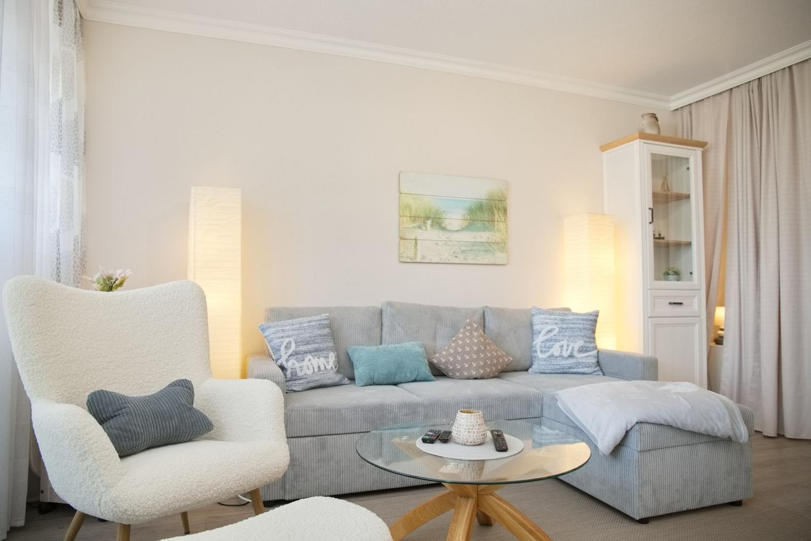 Wohnzimmer mit grauem Sofa, weißen Sessel und Glas-Tisch.