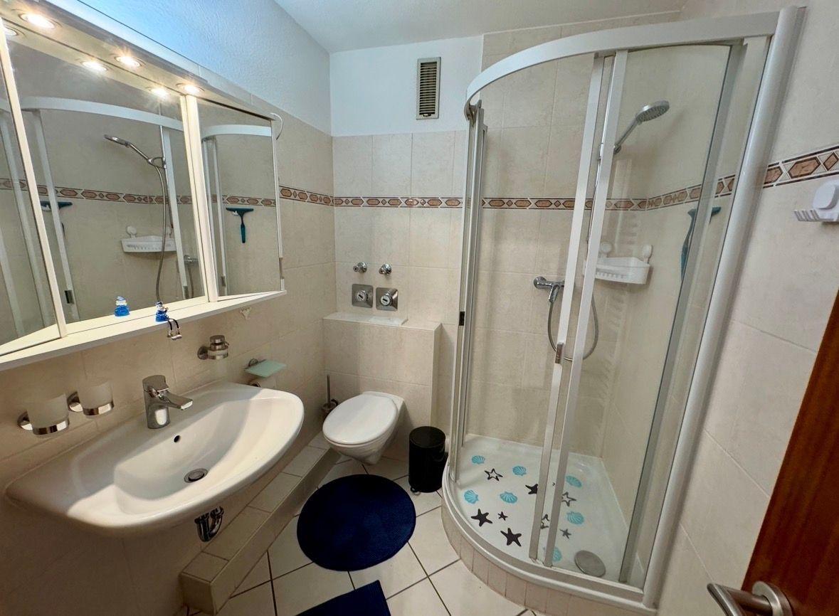 Badezimmer mit Dusche, Waschbecken und Toilette. Fliesen und Spiegel sichtbar.