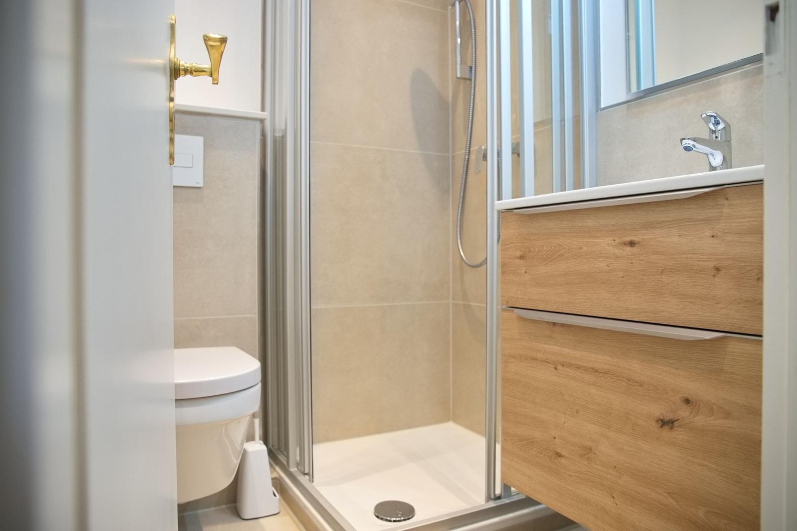 Badezimmer mit Dusche, Waschbecken und Toilette. Holzwaschtisch und goldener Türknauf.