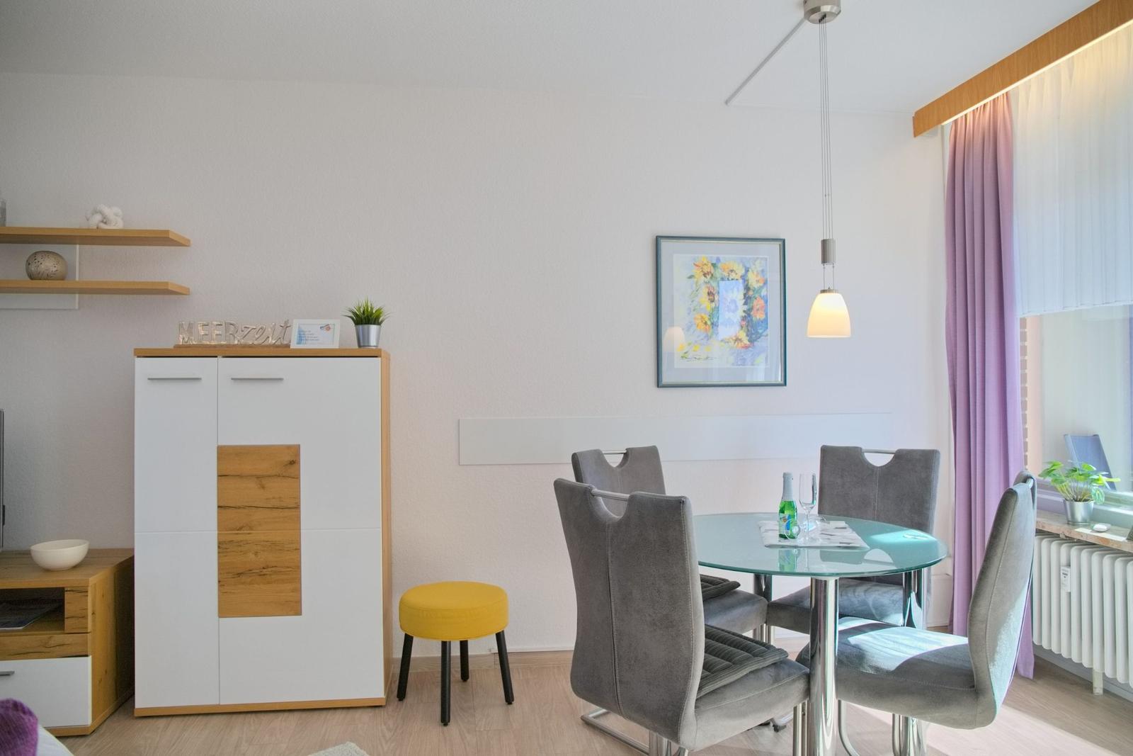 Esszimmer mit Glasrundtisch, vier Stühlen und Schrank. Fenster mit Vorhängen.