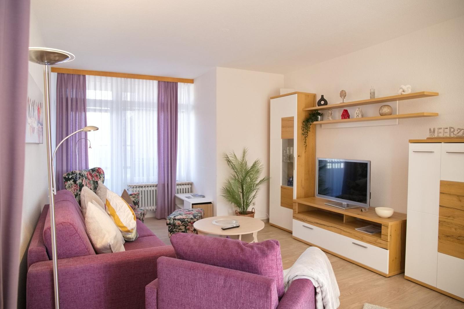 Wohnzimmer mit Sofa, Fernseher und Holzmöbeln. Fenster mit lila Vorhängen.