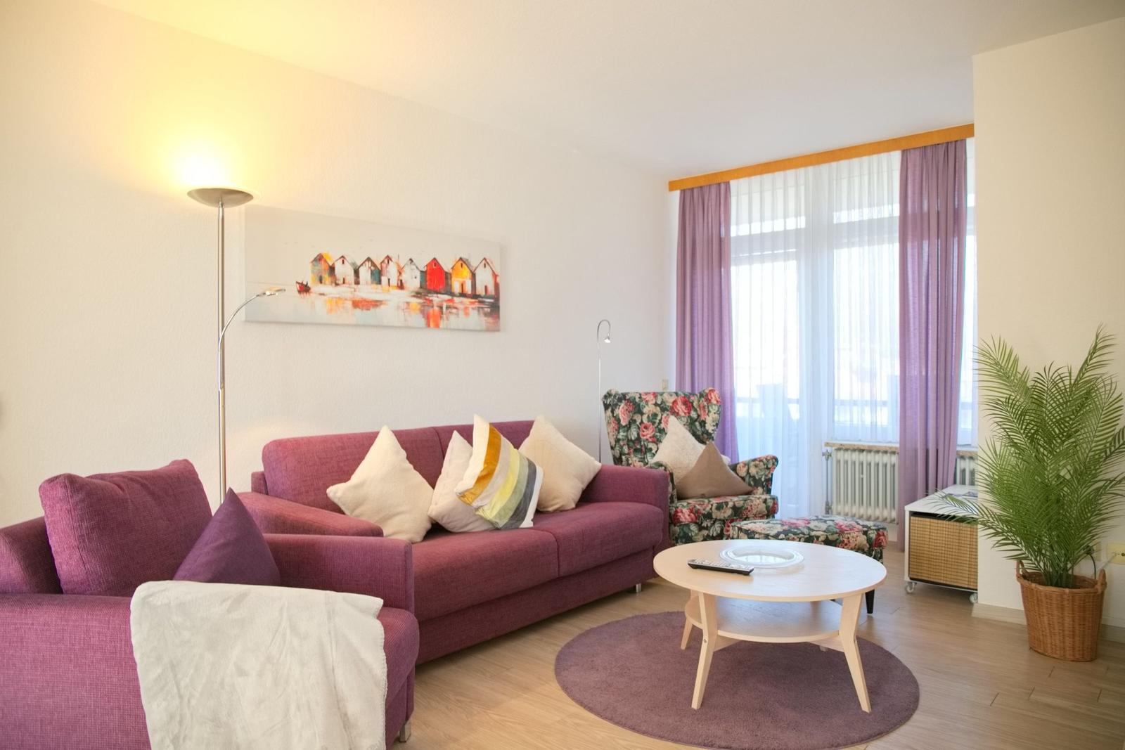 Ein Wohnzimmer mit einem lila Sofa, einem runden Tisch und einem Fenster mit Vorhängen.