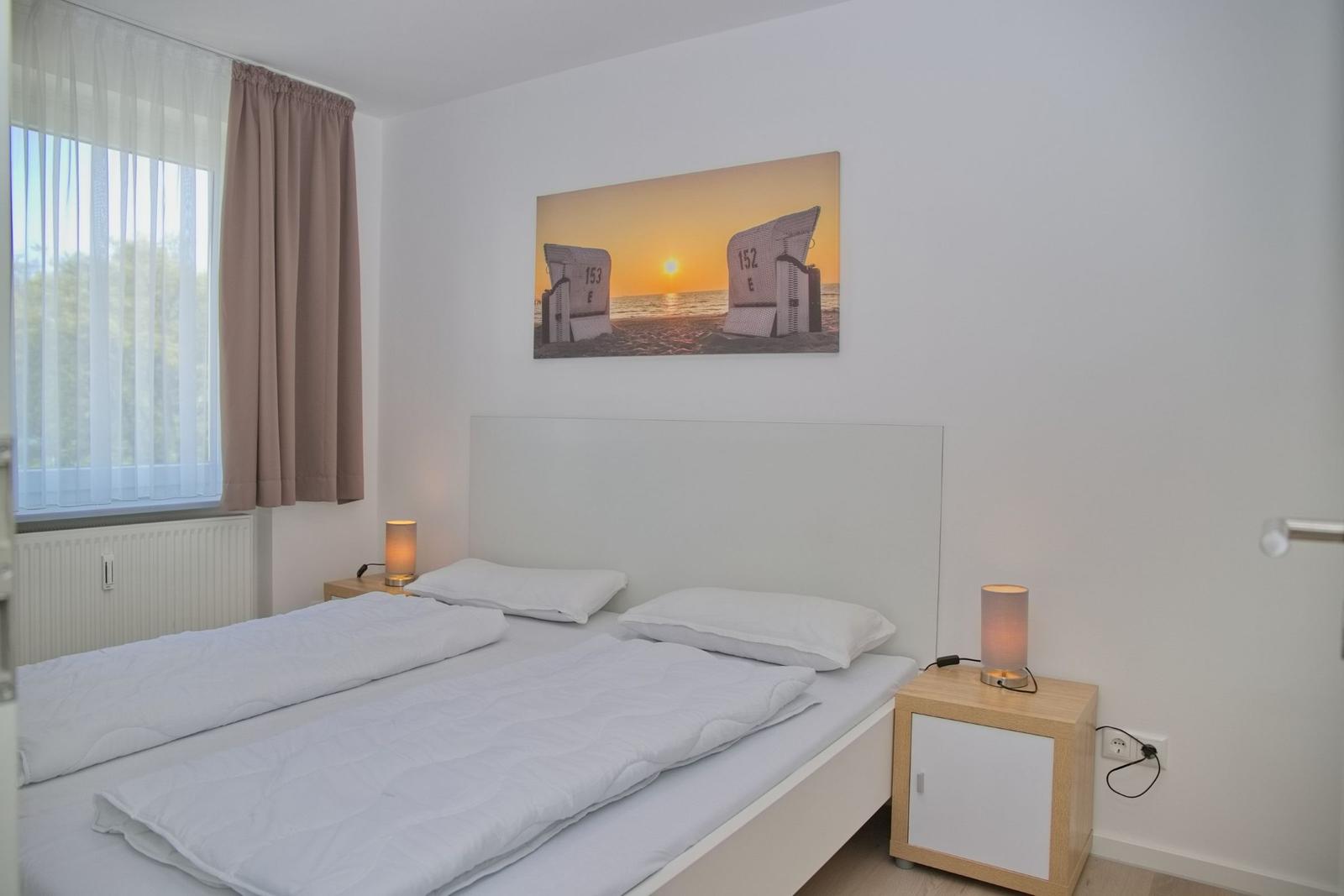 Ein Schlafzimmer mit Doppelbett, Nachttischen und Fenster. Wandbild zeigt Strandkörbe.
