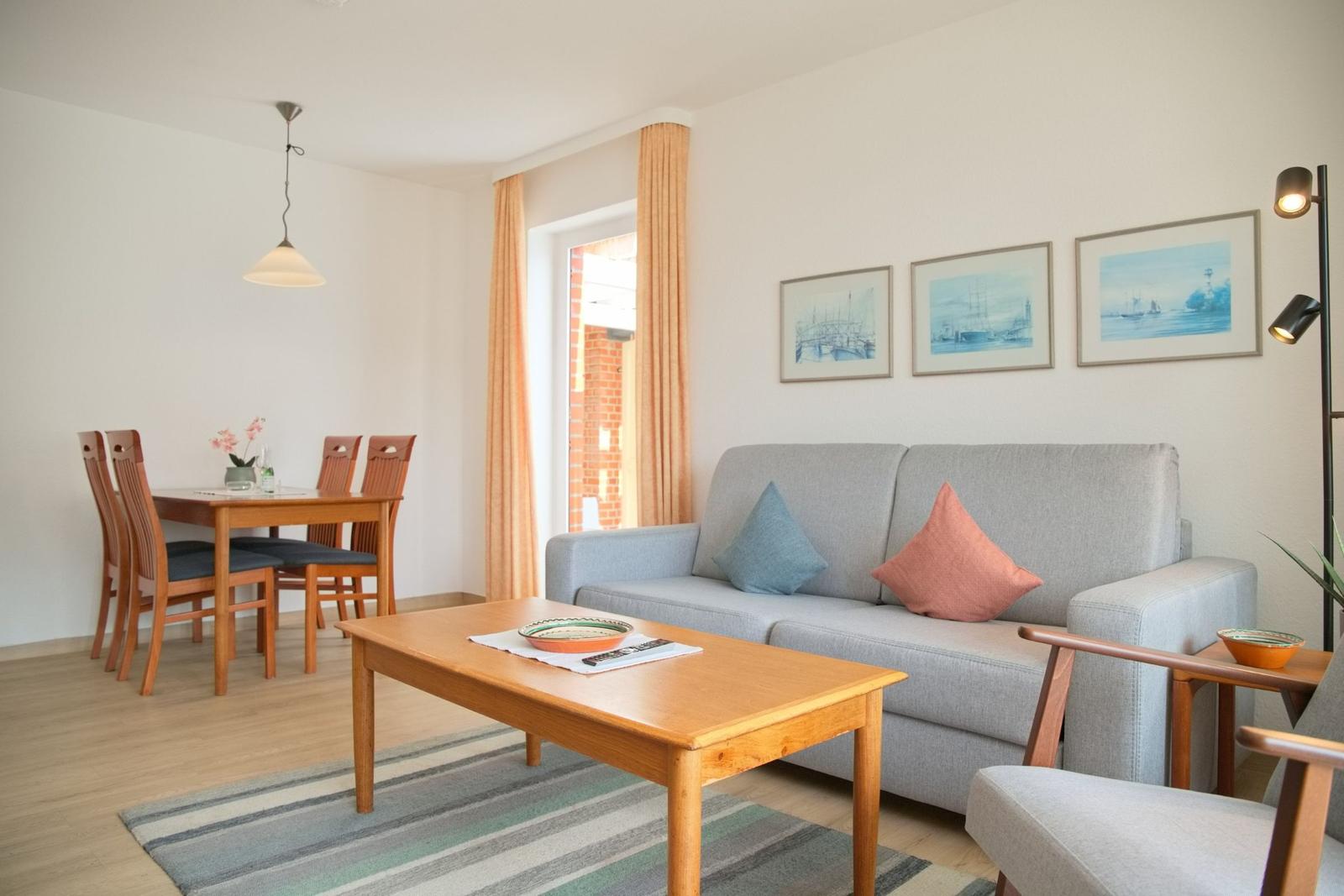 Wohn- und Essbereich mit Sofa, Tisch und Stühlen. Fenster mit Vorhängen. Wandbilder.