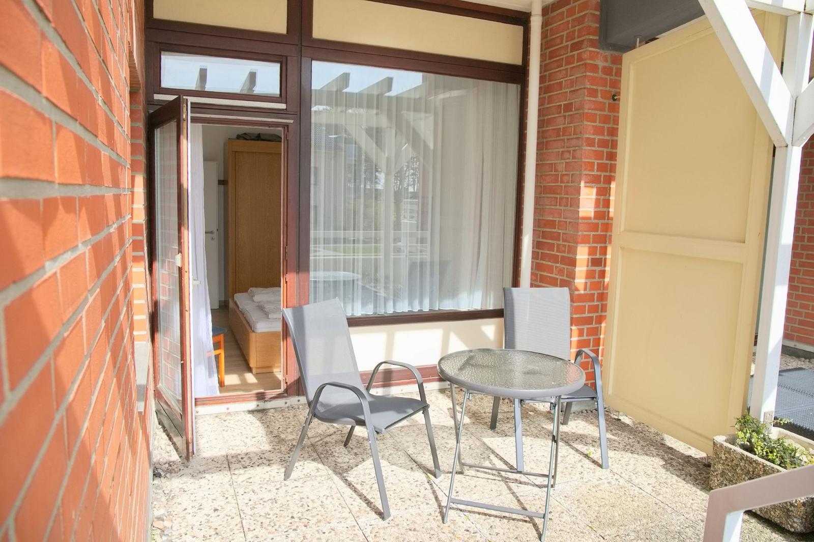 Terrasse mit Tisch und zwei Stühlen. Durch die Tür ist ein Schlafzimmer sichtbar.