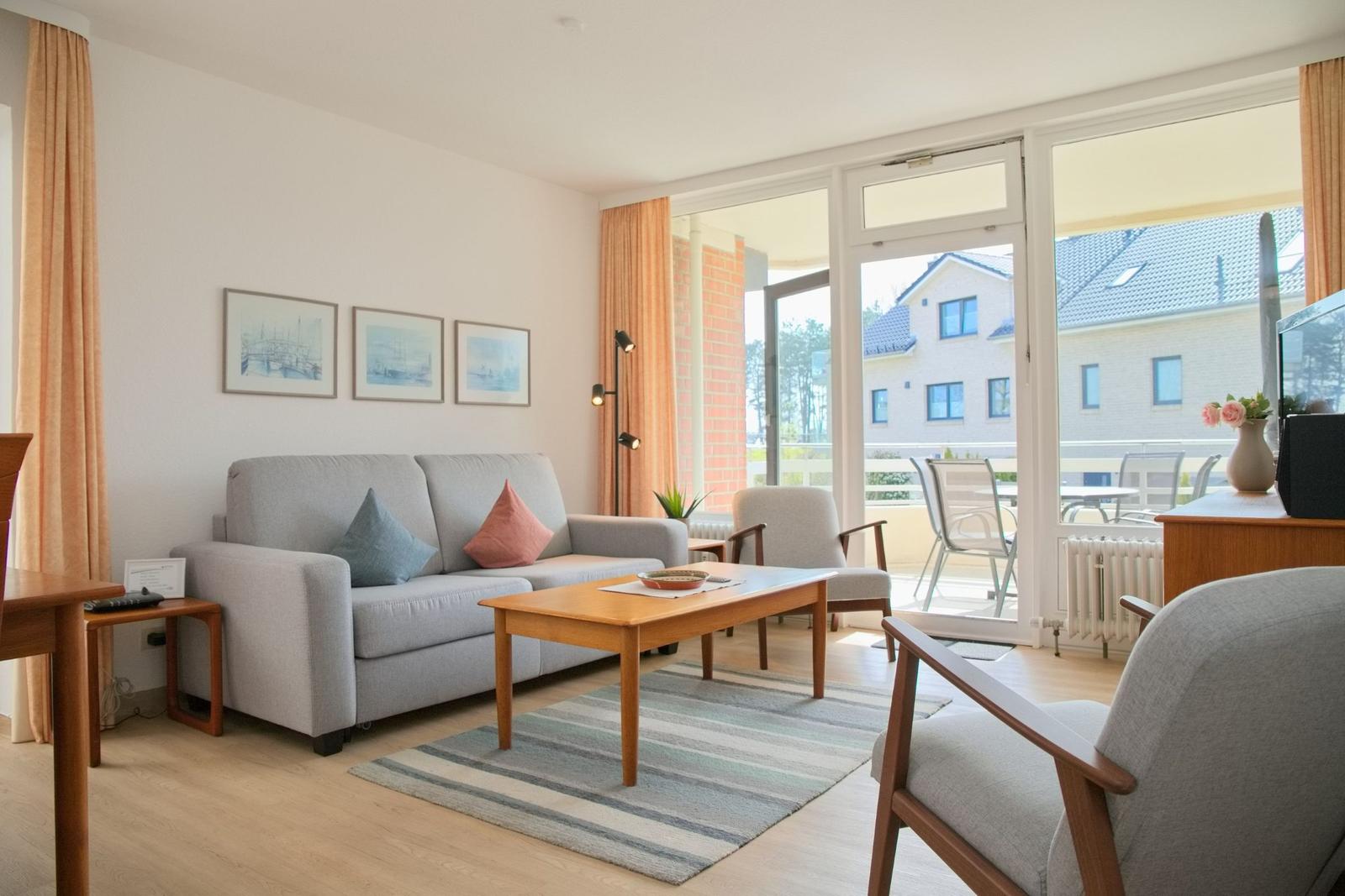 Wohnzimmer mit Sofa, Tisch und Blick auf Balkon. An der Wand hängen drei Bilder.