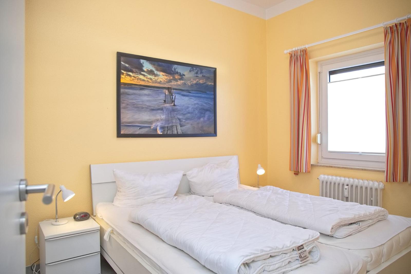 Schlafzimmer mit Doppelbett, Fenster und Wandbild. Gelbe Wände und weiße Möbel.