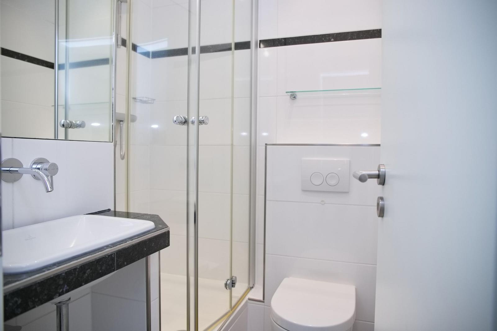 Badezimmer mit Dusche, Waschbecken und Toilette. Weiße Fliesen und Glaswand.