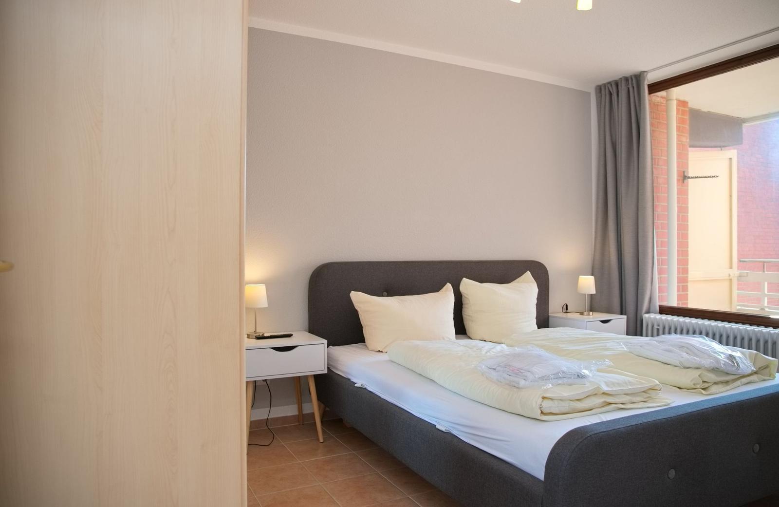 Schlafzimmer mit Doppelbett, Nachttischen und Fensterblick auf Balkon.