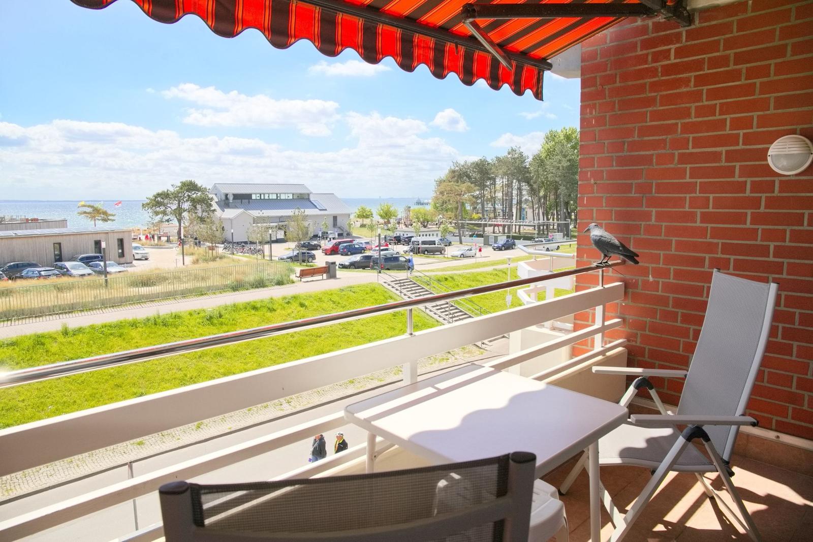 Terrasse mit Tisch, Stühlen und rotem Markisen, Blick auf Grünfläche und Strand.