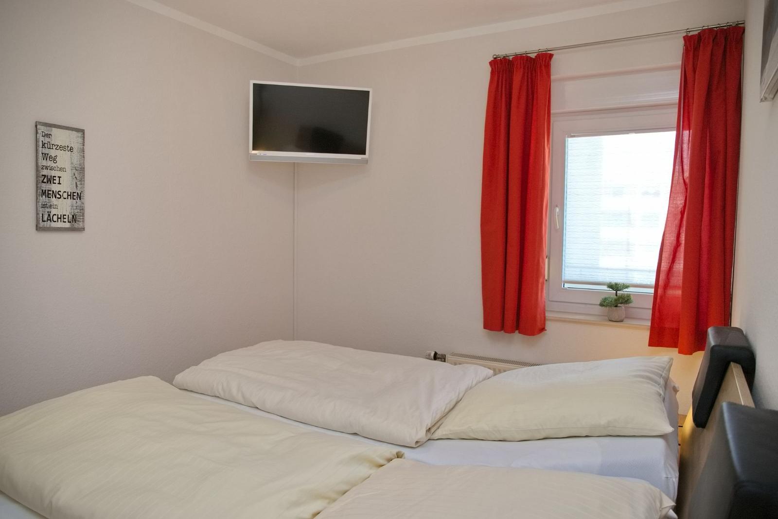 Zimmer mit Doppelbett, Fernseher und Fenster mit roten Vorhängen.