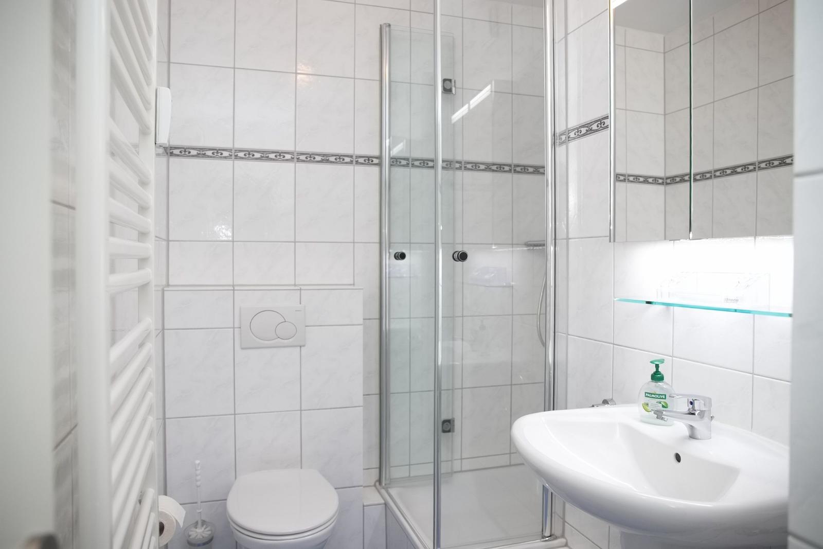 Badezimmer mit Dusche, Waschbecken und Toilette. Weiße Fliesen und Spiegelschrank.