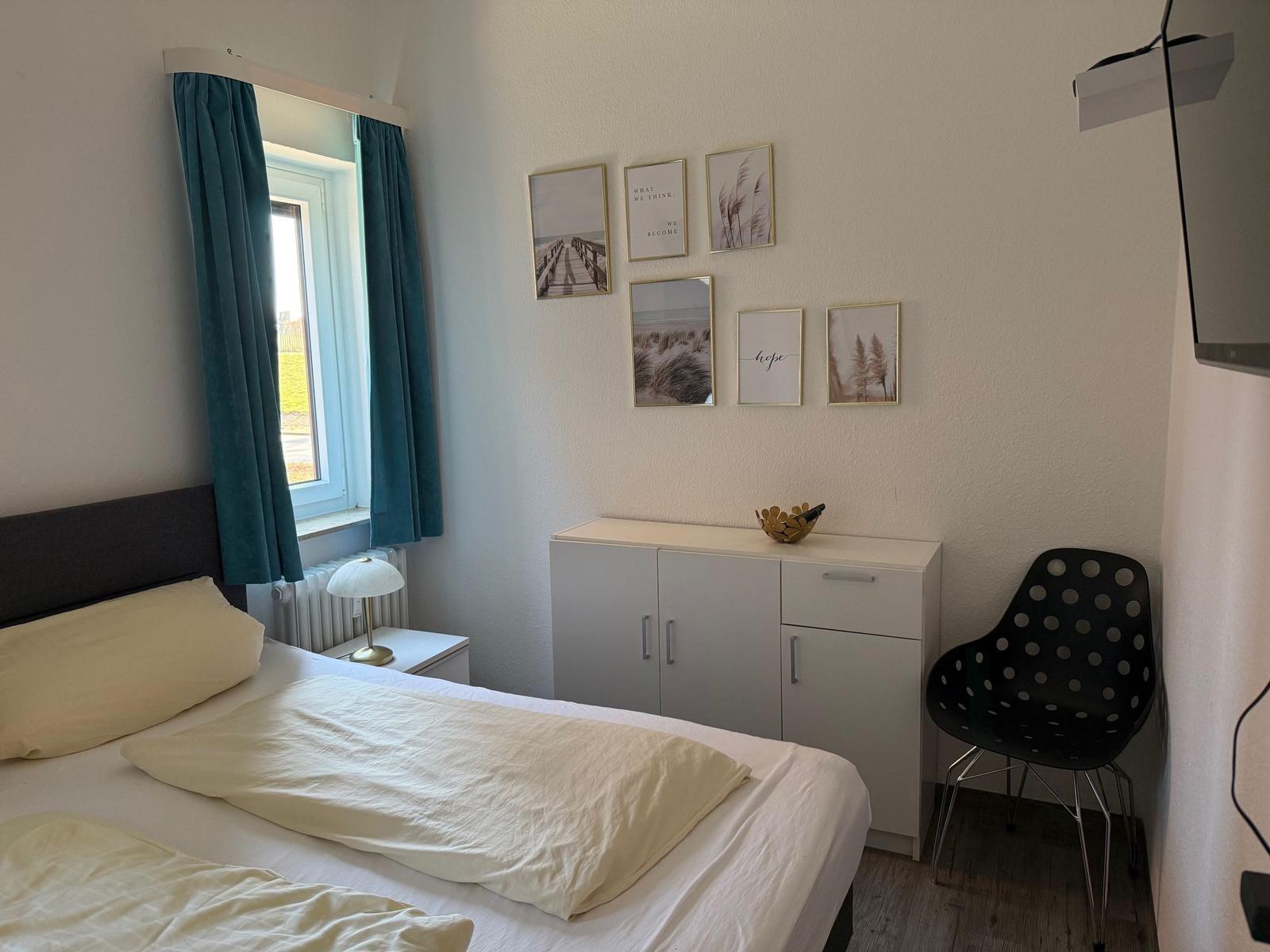 Schlafzimmer mit Bett, Schrank, Stuhl und Fenster mit Vorhängen.