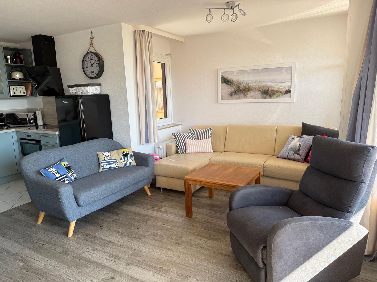 Wohnzimmer mit Sofa, Sessel und Küchenbereich. Wanduhr und Strandbild an der Wand.