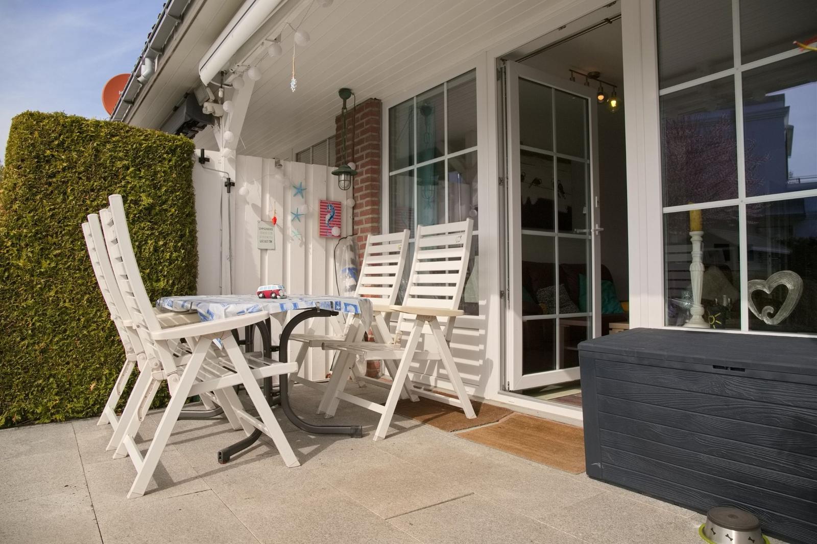 Terrasse mit weißem Tisch und Stühlen, Sicht auf Wohnzimmer durch Glastüren.