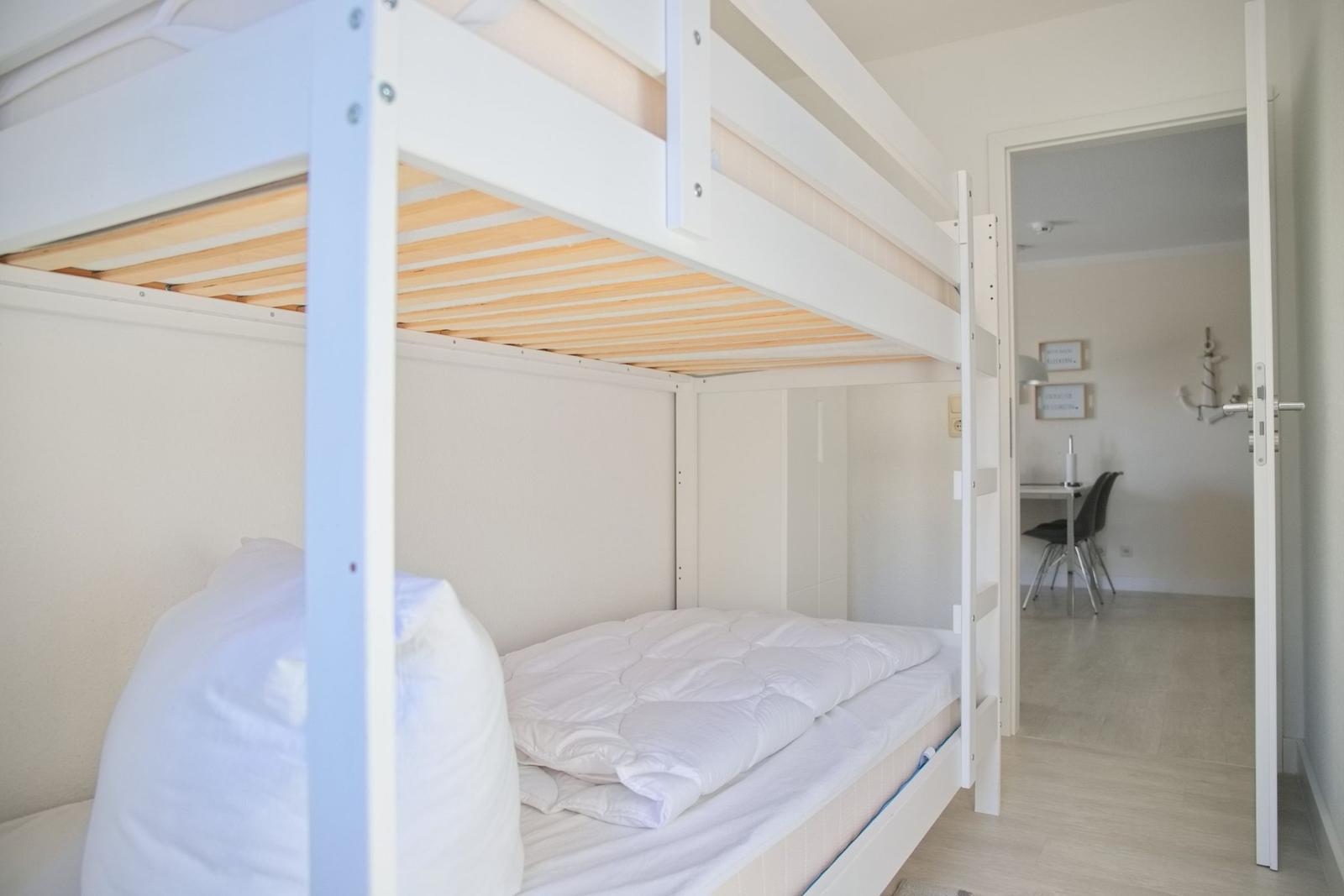 Doppelbett mit Matratze und Kissen. Durchgang zur Küche mit Tisch und Stühlen.