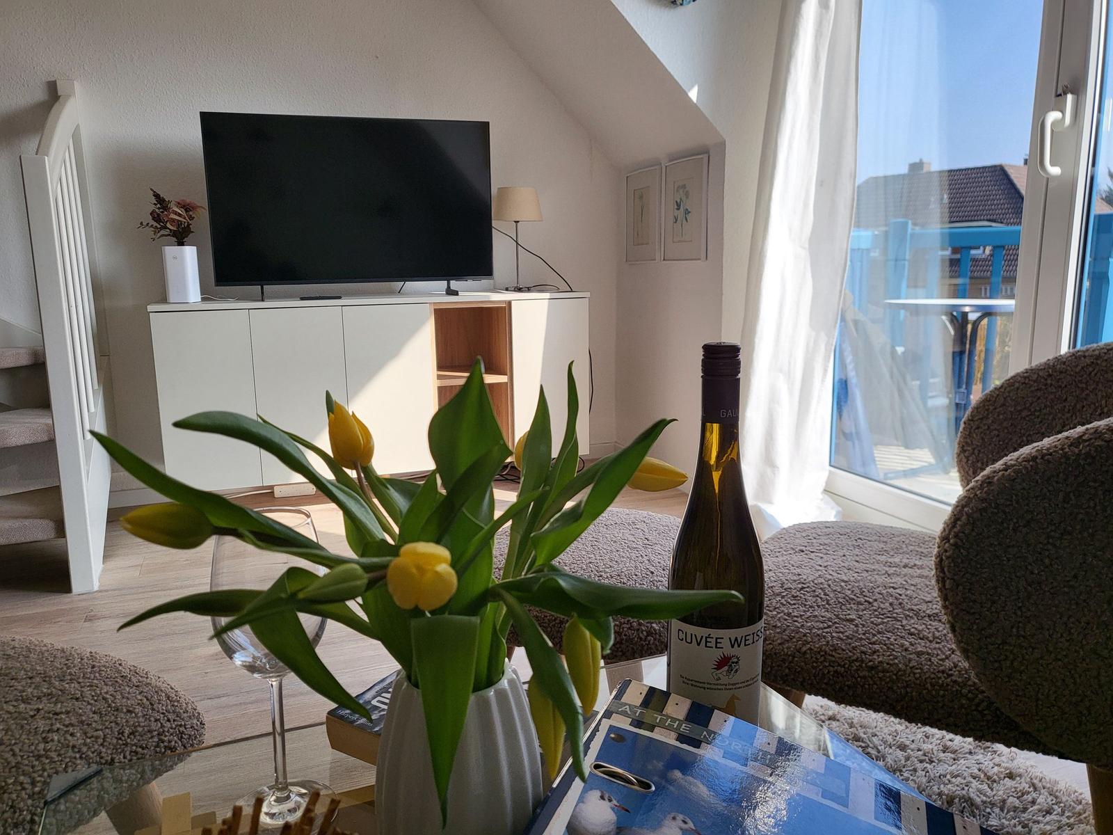 Wohnzimmer mit Fernseher, Blumen und Blick auf Balkon.