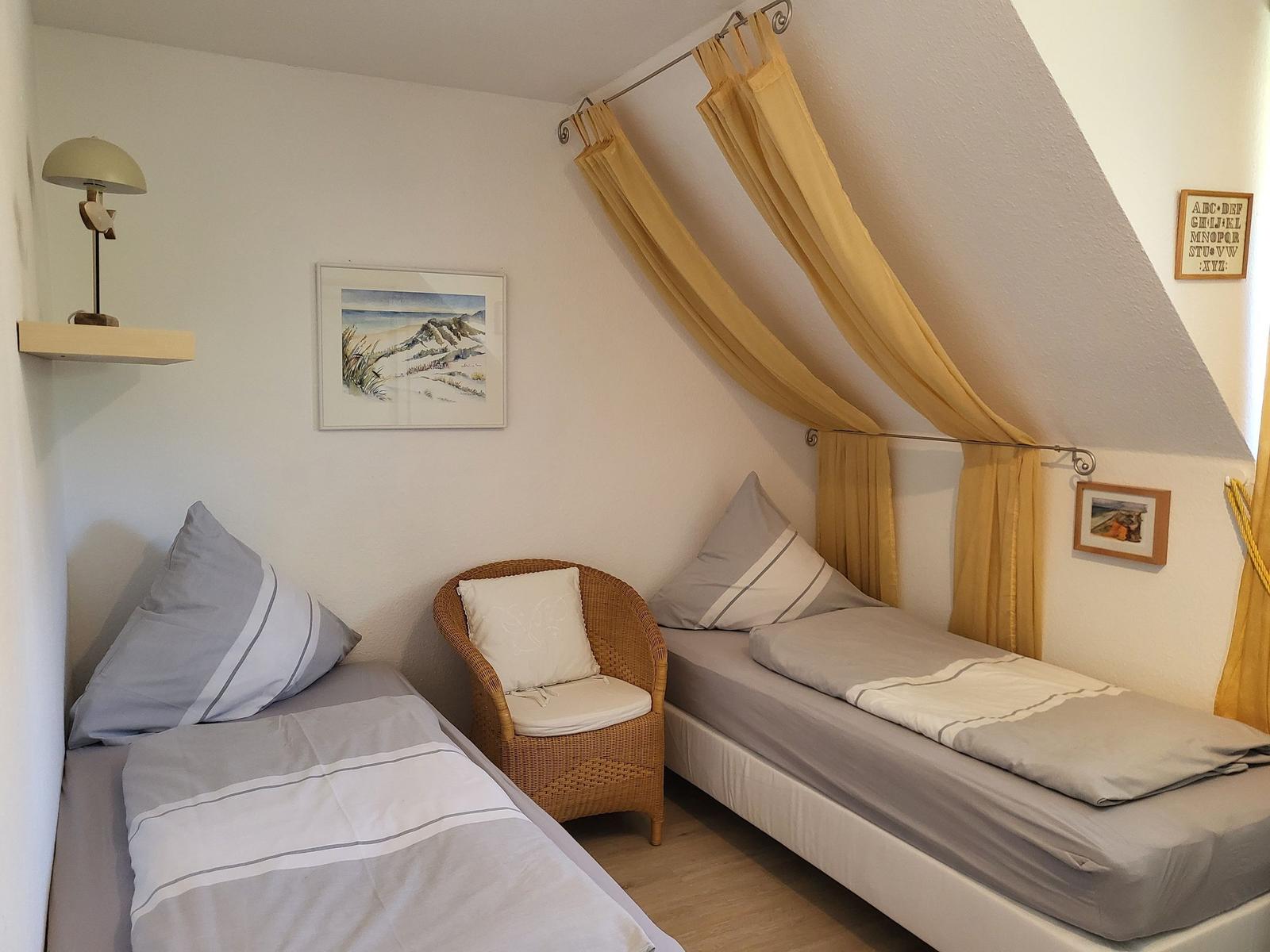 Zimmer mit zwei Betten, Stuhl und Gardinen unter Dachschrägen.