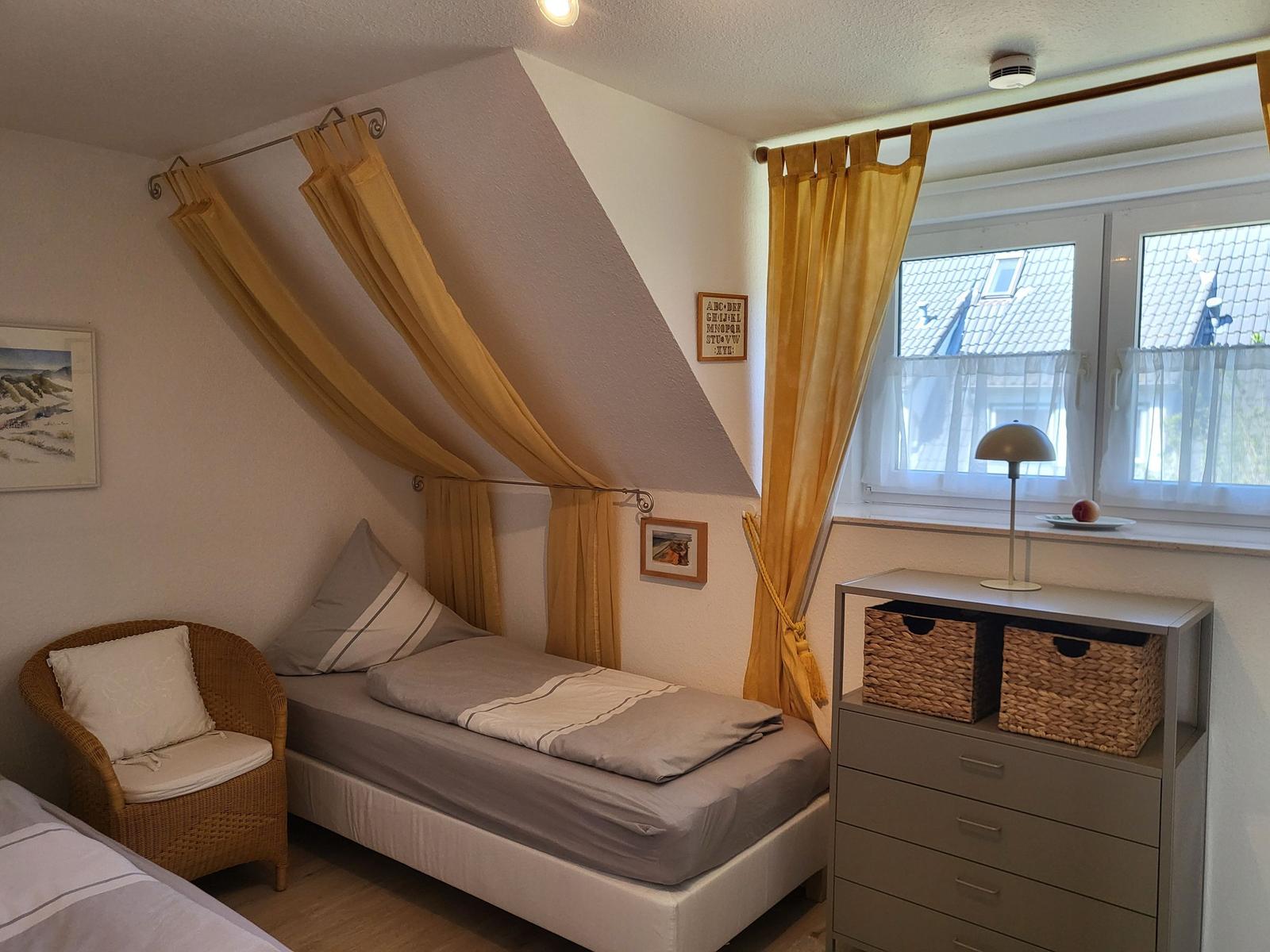 Schlafzimmer mit Dachschrägen, Bett, Stuhl und Schrank. Fenster mit Vorhängen.