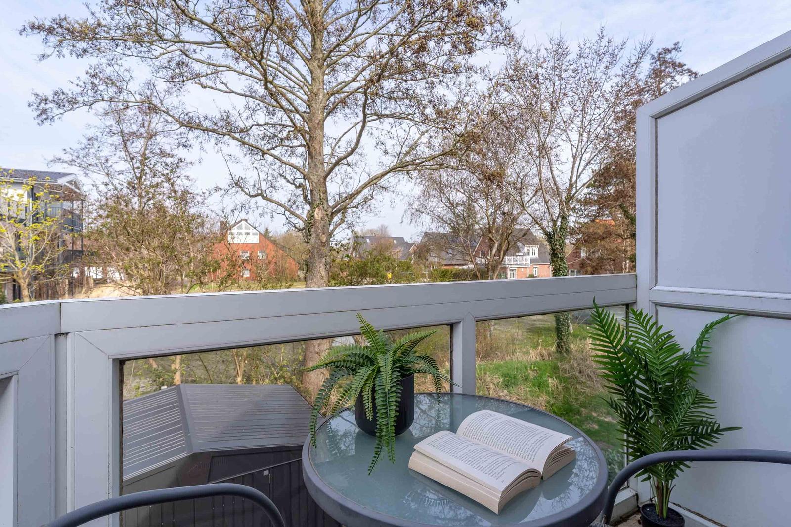 Terrasse mit Tisch, Pflanzen und offenem Buch. Blick auf Bäume und Häuser.