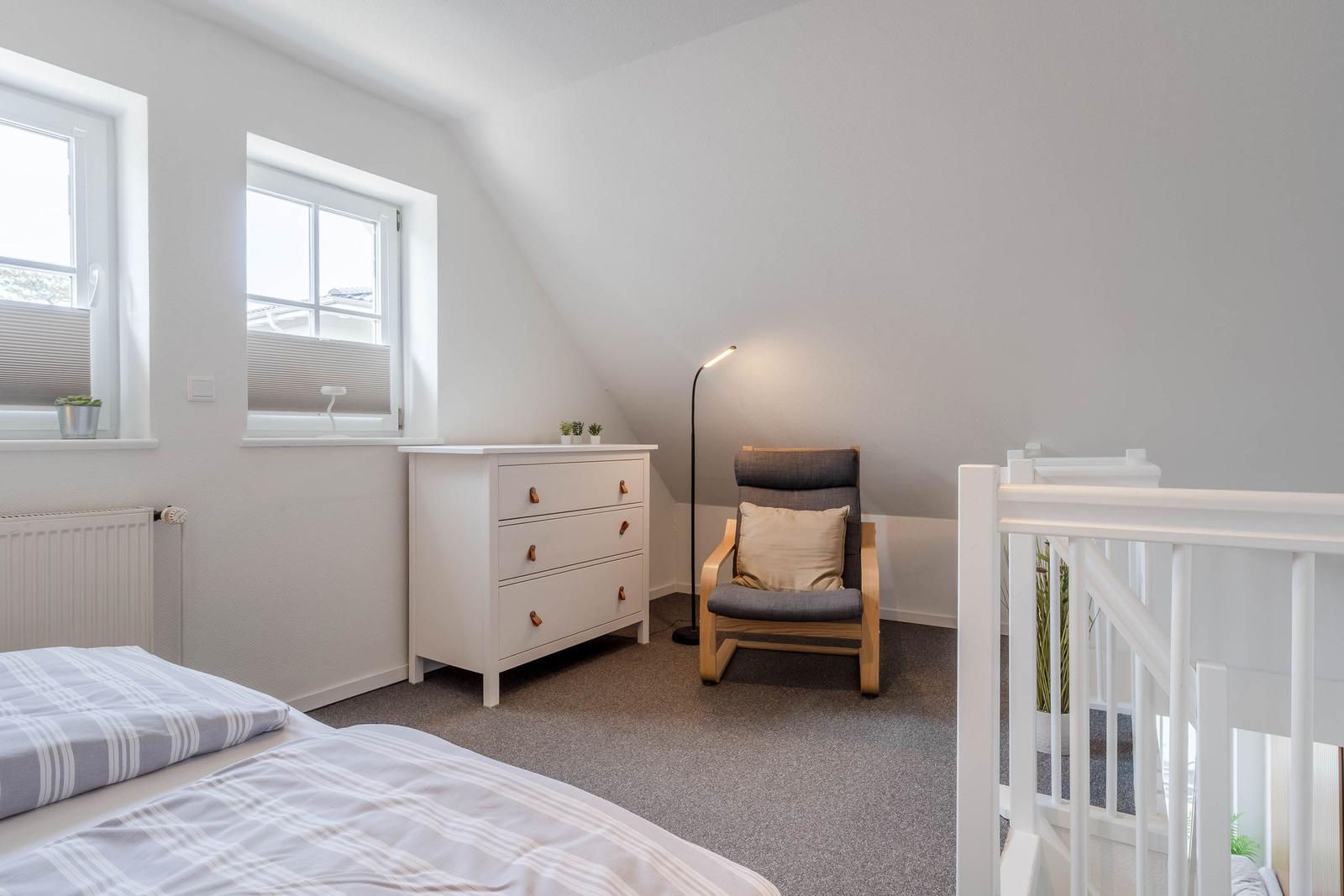 Schlafzimmer mit Bett, Schrank, Sessel und zwei Fenstern.