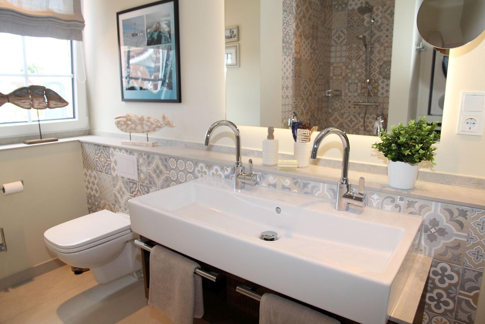 Badezimmer mit Waschtisch, Spiegel und Dusche. Wandfliesen und Pflanze sind sichtbar.