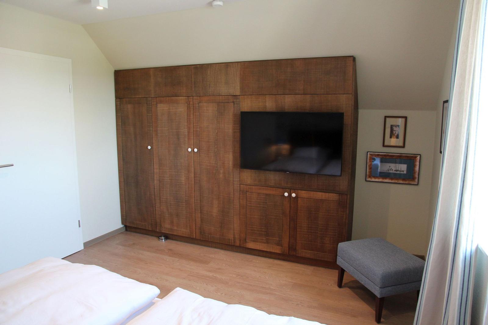 Schlafzimmer mit Holzschrank, Fernseher und grauem Hocker.