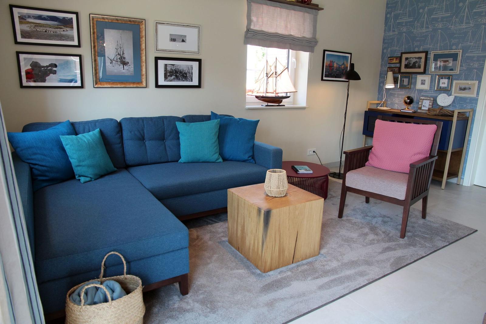 Ein Wohnzimmer mit blauem Sofa, Holztisch und rosa Sessel. Wand mit Schiffsmotiven und Bildern.