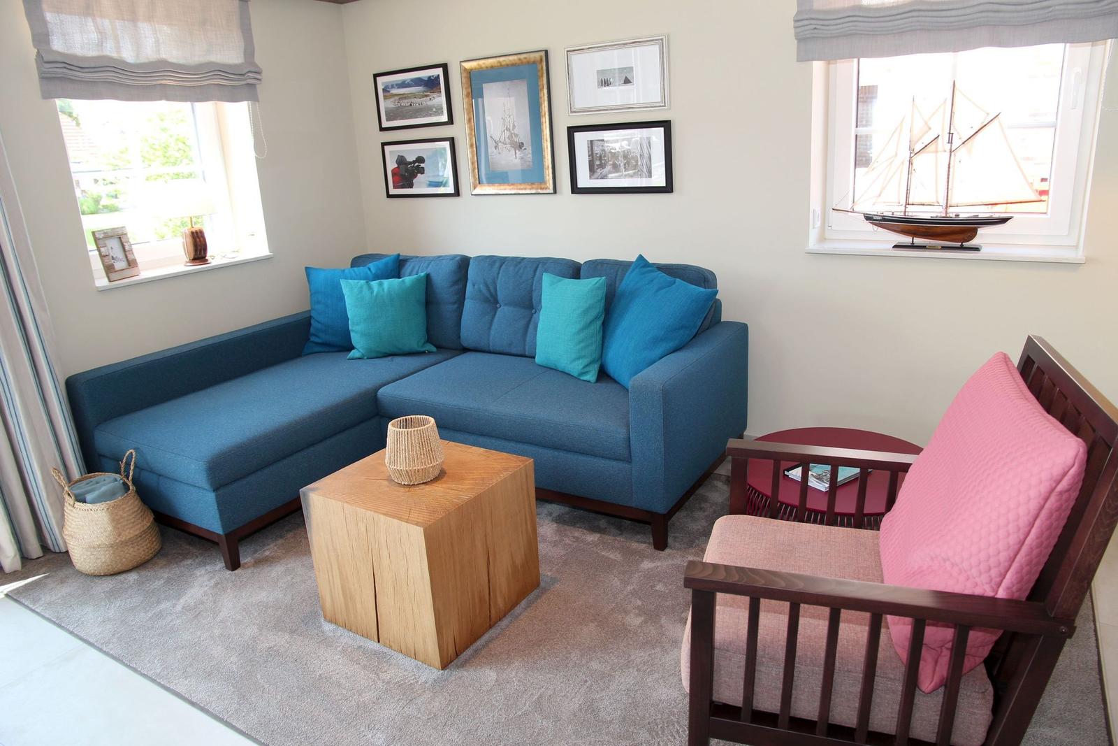 Ein Wohnzimmer mit blauem Sofa, rosa Sessel und Holztisch. An den Wänden hängen Bilder.