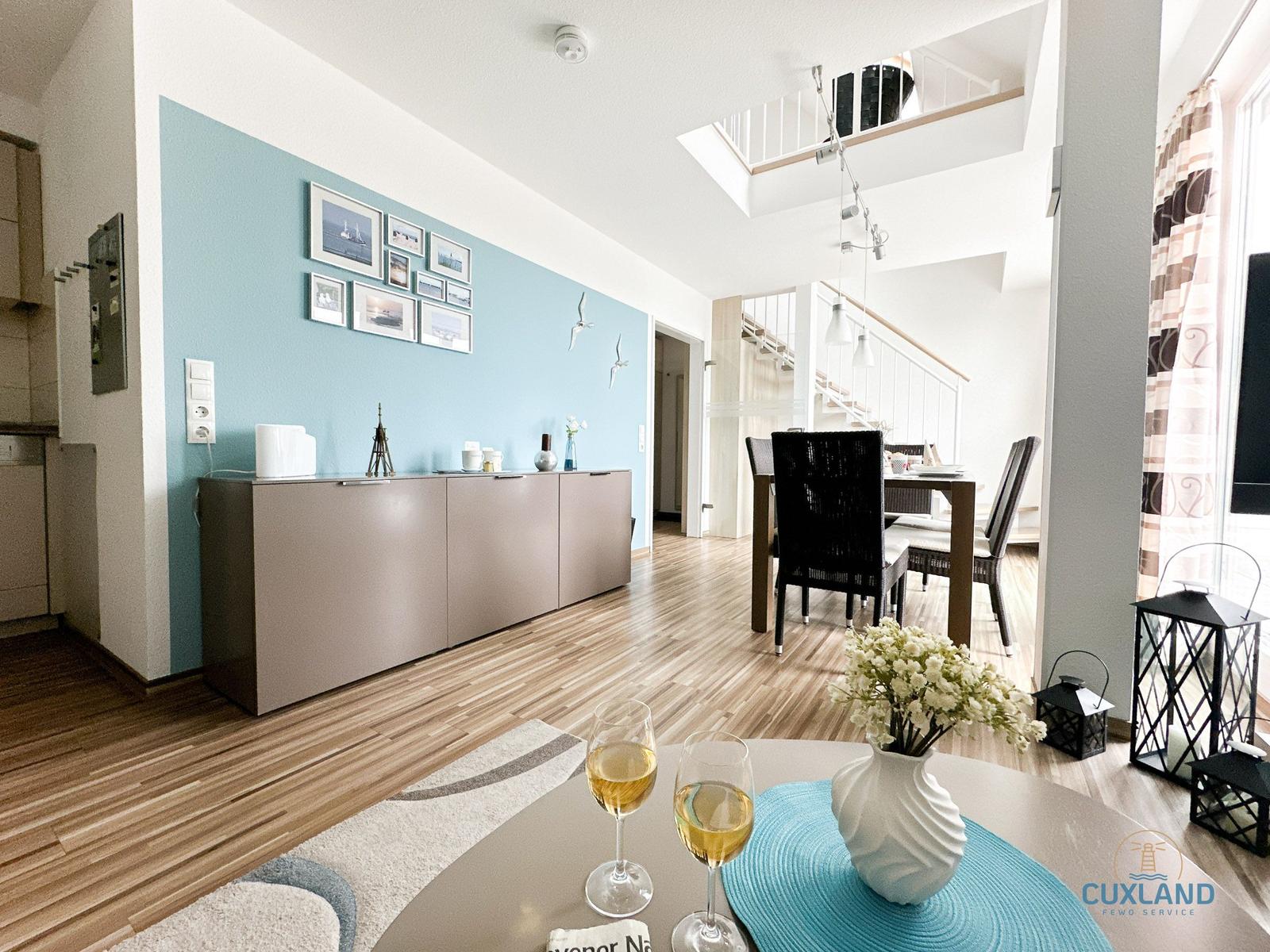 Wohnbereich mit Essbereich, blauer Wand und Holzboden. Tisch mit Weingläsern und Blumen.