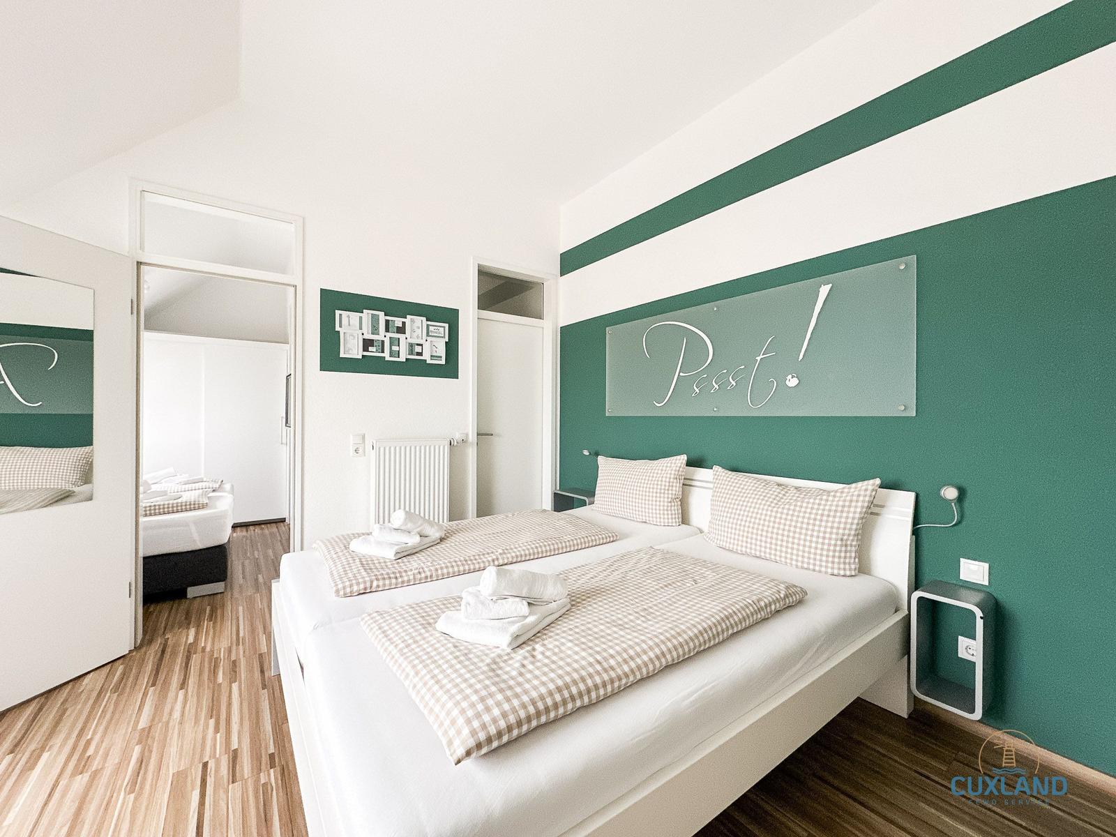 Schlafzimmer mit Doppelbett, grüner Wand und Spiegel. Flur und weitere Zimmer sichtbar.
